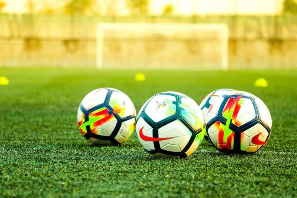Three bright soccer balls sit on a green field with a goal in the blurred background.