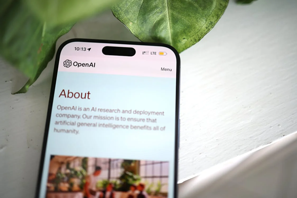 OpenAI and Microsoft End their Business Agreement Close-up of a smartphone screen showing the OpenAI About page with a pastel gradient header and a plant in the background.