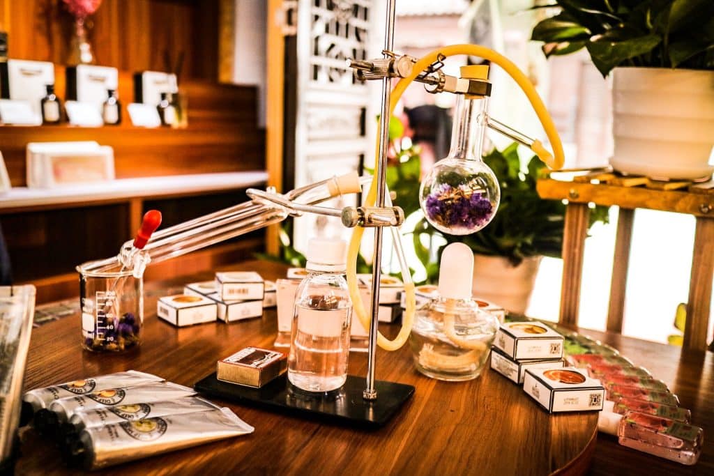 Men’s Fragrances That Will Make You Discover the Charm of Artistic Perfumery Lab-style setup on a wooden table with glass flasks, tubing, and a round-bottom flask containing purple flowers