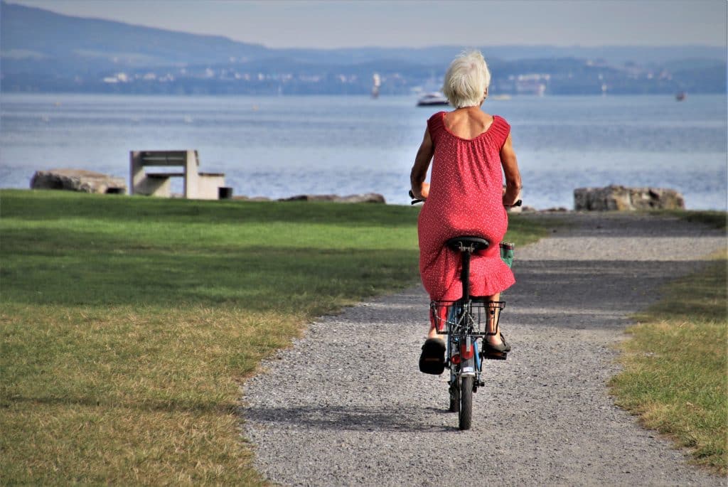 How to Maintain Your Active Lifestyle Long into Your Golden Years Older woman in a red dress rides a bicycle along a gravel path beside a lake, viewed from behind with green grass and distant hills.