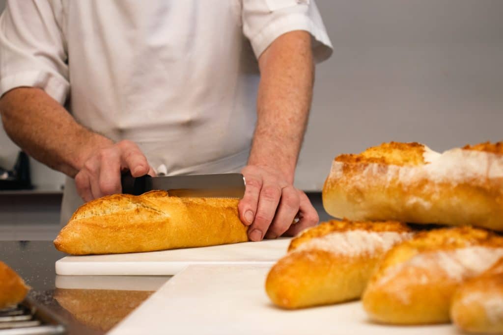 Materials Matter: Breathable, Stain Resistant and Sustainable Fabrics Used in Chef Jackets Baker slices a crusty baguette on a white cutting board with more baguettes nearby in a bakery kitchen