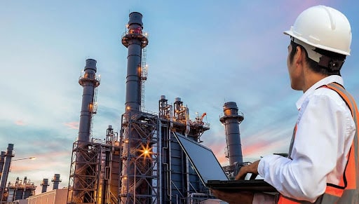 Engineer in a hard hat and orange safety vest reviews a tablet while standing at a large refinery complex with tall chimneys at sunset.