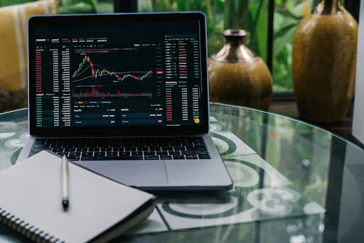 Living In Australia: A Guide To Investment Opportunities For New Migrants Laptop shows a stock trading dashboard with charts and data on a glass outdoor table, with a notebook nearby.