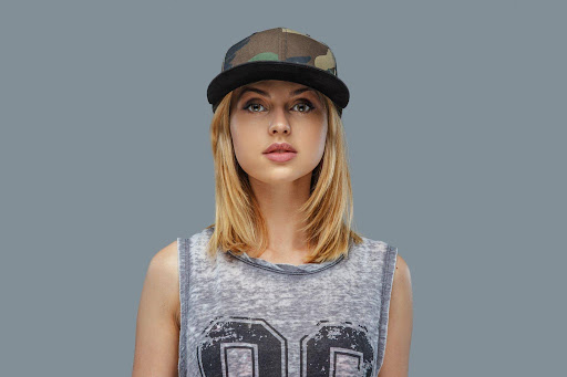 The Rise of Smart Streetwear: How Technology Is Redefining Modern Hat Culture Portrait of a young woman wearing a camouflage cap and a gray sleeveless graphic tee, facing the camera against a gray background.