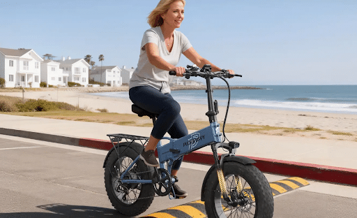 How to Choose the Best Ebikes for Sale in 2026 Woman rides a blue compact electric bike along a sunny seaside street with houses and the beach in the background.