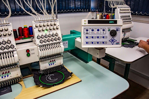 Are Polyester T Shirts Good for Printing and Embroidery? Industrial embroidery machine setup with multiple thread spools and digital control panel, hooping a black fabric sample on a sewing bed.