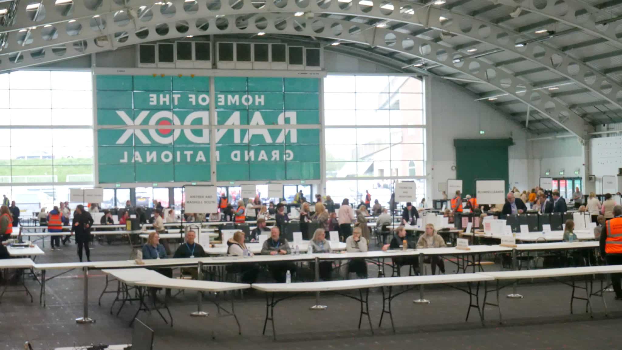 Busy indoor registration area with many people at long white tables, signs, and volunteers in orange vests; teal banner on the back wall remains visible