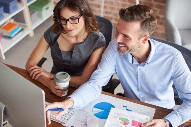 Two coworkers collaborate at a desk, reviewing data on a computer with charts and a coffee cup nearby on a modern office table.Changes and insights are being discussed.