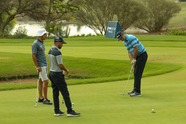 Three golfers on a putting green; one in a blue striped shirt lines up a putt while two others watch.