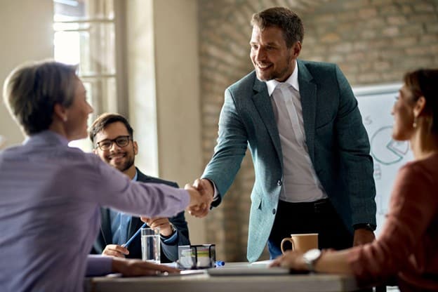 Two colleagues shake hands across a conference table while others look on with smiles in a bright office.