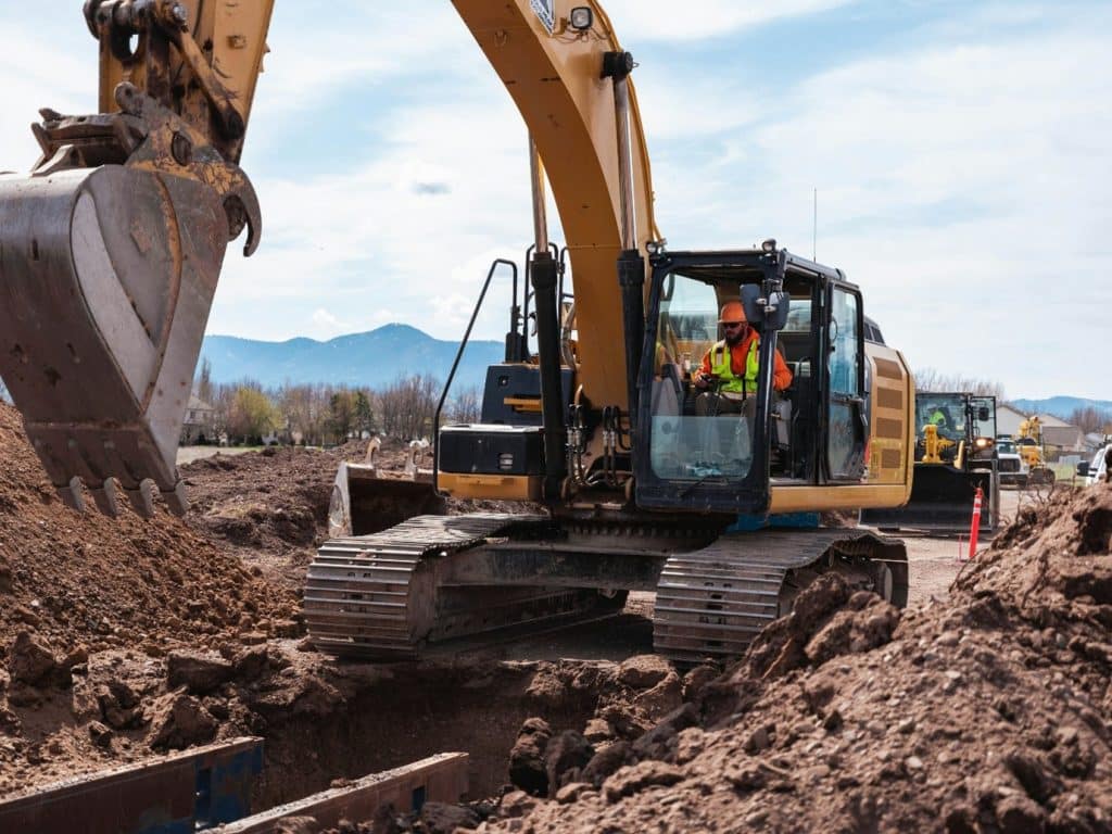Yellow excavator at a construction site digging a trench, worker in a safety vest inside the cab with a mountainous backdrop.”] ,
