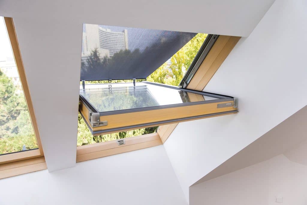 Fresh Air in Your Loft: Ventilation Benefits of Centre Pivot Windows Open skylight window in a slanted white ceiling with wood trim, letting in daylight and a view of trees outside.