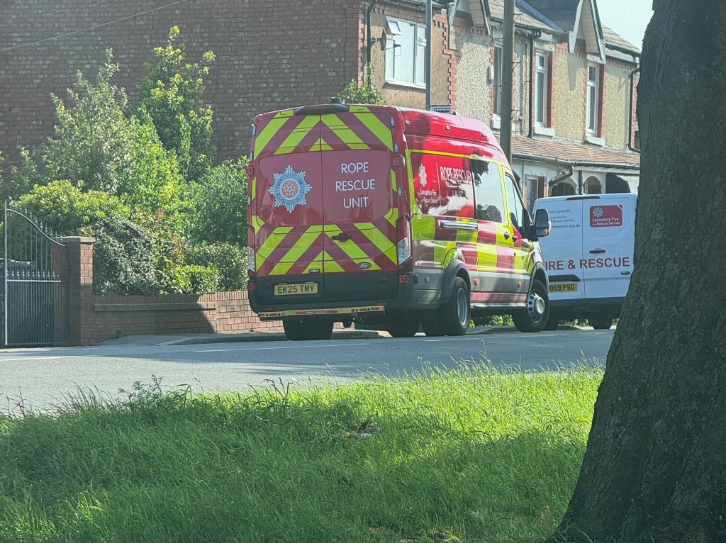 Multiple emergency crews attend incident in Banks Red Lancashire Fire and Rescue rope rescue unit van parked on a residential street with brick houses and greenery in the background.