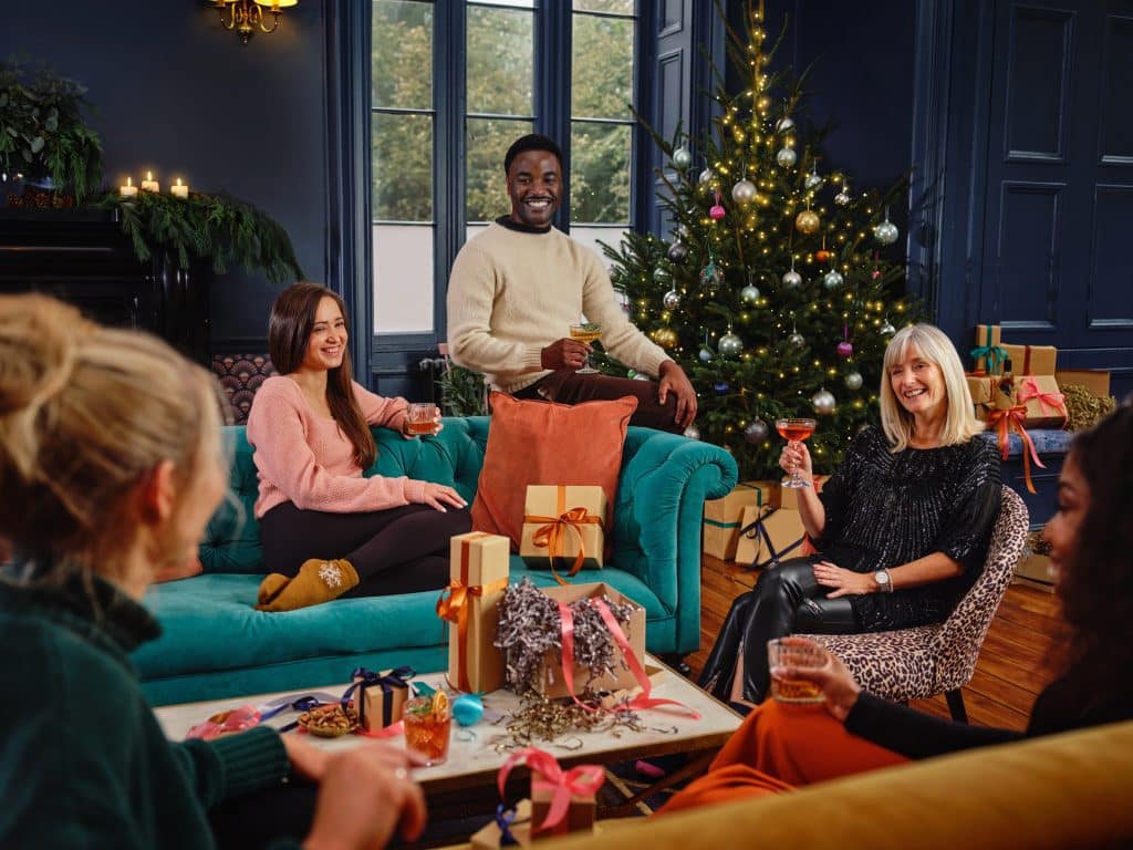 Physical gift cards remain popular as shoppers look for simple and flexible presents Group of friends gathered on teal couches around a decorated Christmas tree, sharing drinks and gifts in a festive living room.