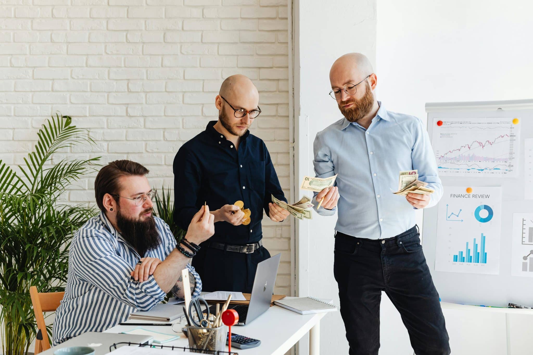 Three men in a modern office: one seated at a desk with a laptop, another standing and counting cash, a third holding coins, with charts on a whiteboard in the background.
