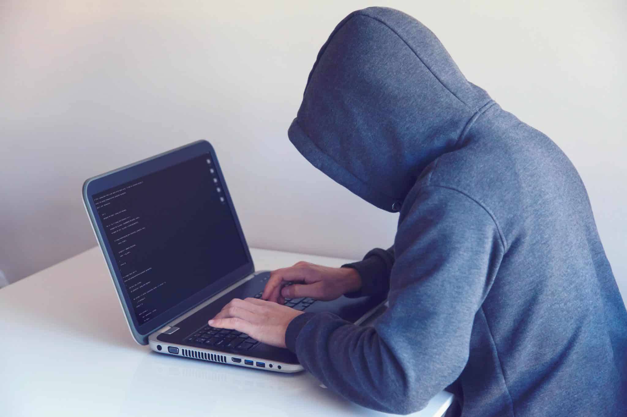 Person in a gray hoodie typing on a laptop showing lines of code on the screen, seated at a white desk.