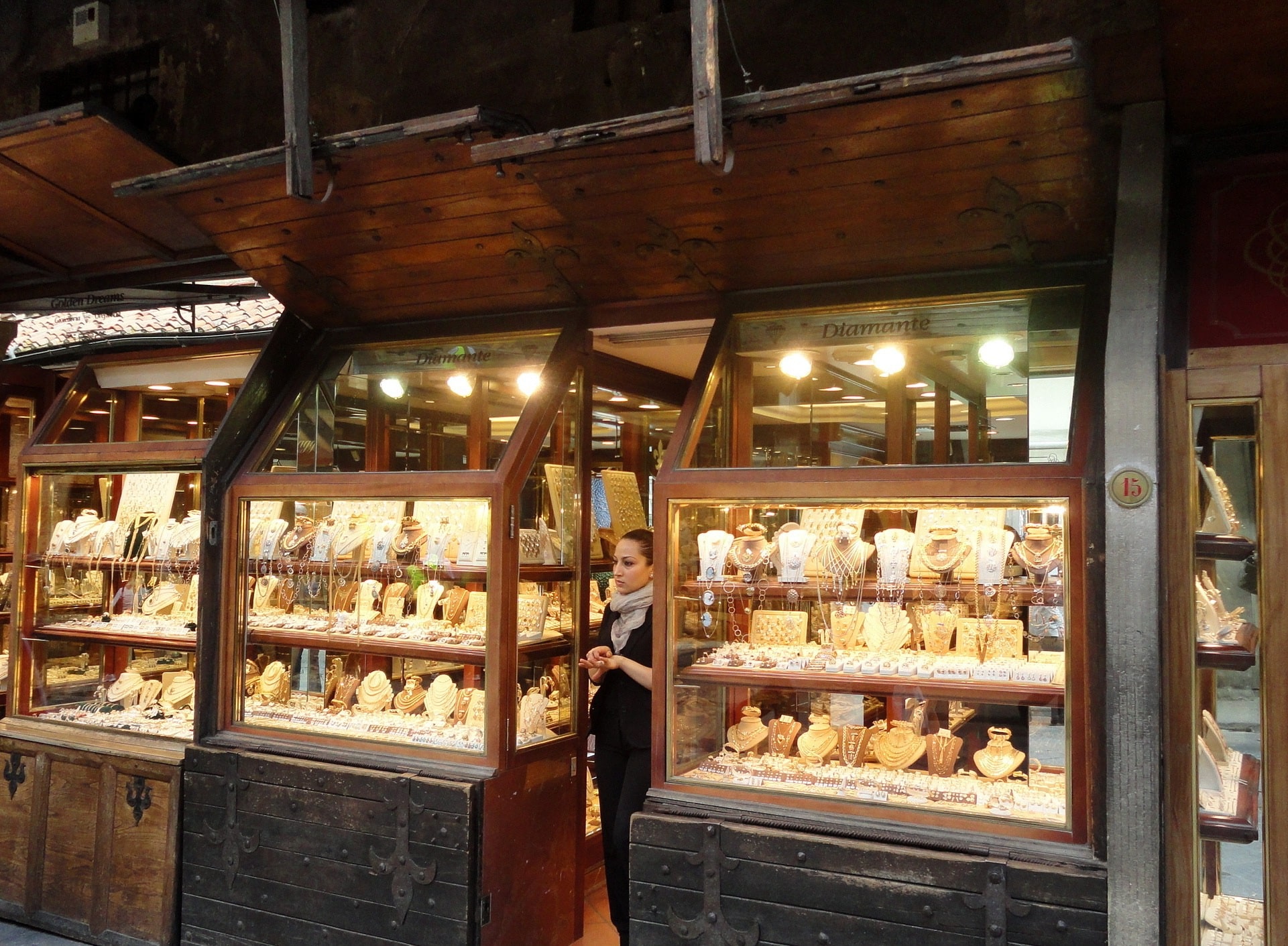 Jewelry storefront at night with glass display cases full of necklaces and bracelets; a woman in black stands at the doorway.