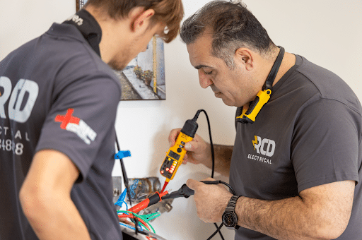 Two electricians in gray shirts, branded RCD Electrical, test and connect wires with a multimeter and pliers near a wall outlet.