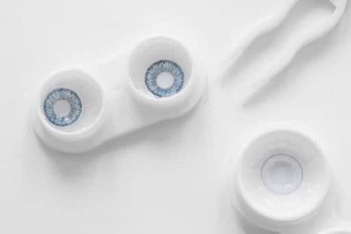 Silicone Hydrogel vs Hydrogel Eye Contact Lenses Two blue contact lenses in open white lens cases on a white surface, with an additional empty case nearby.
