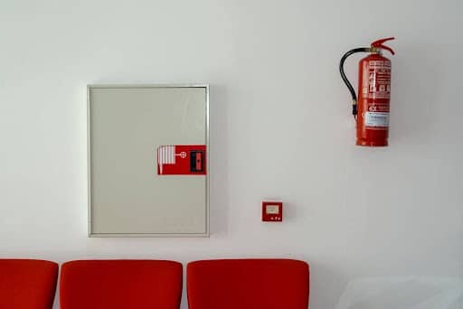 Fire Safety Compliance for UK Small Businesses White wall with a rectangular mirror cabinet, a high red fire extinguisher, a small red emergency switch, and red chairs along the bottom.