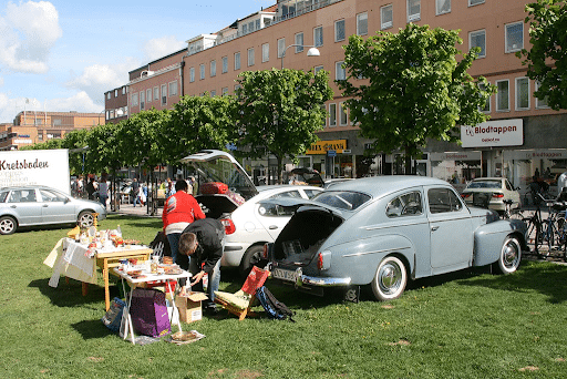 Car Boot Sale Tips That Actually Work in 2026: A Practical Guide for First-Time Sellers Vintage blue car with its trunk open at an outdoor street market on grass; shoppers browse tables of goods with vendors nearby, brick buildings in the background.