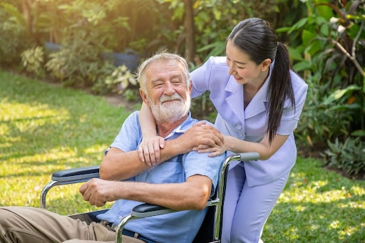 How to Choose the Right Elderly Caregiver for Your Loved One Caregiver in lavender scrubs gently hugs elderly man in a wheelchair in a sunny garden.