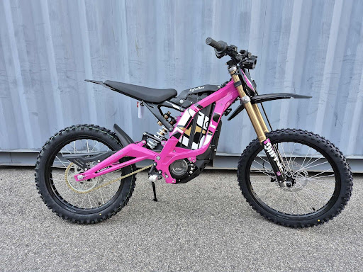 Can Electric Dirt Bikes Replace Traditional Gas-Powered Bikes? Pink full-suspension dirt bike with gold front forks and knobby tires parked on pavement in front of a blue wall.