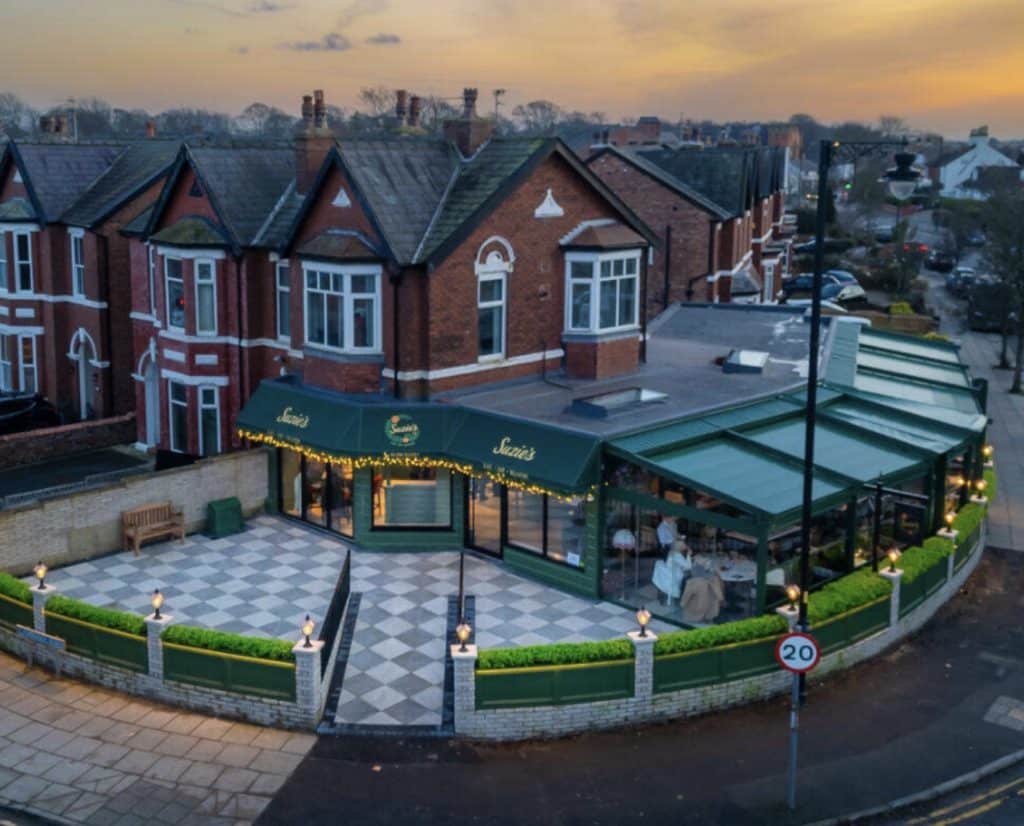 Popular Churchtown eatery launch appeal to save much loved terrace A street-side cafe called Suzie's with a green awning, outdoor seating, and string lights next to red brick houses at sunset.