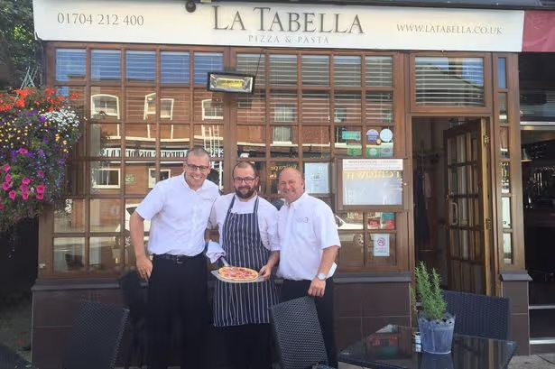 Three men stand outside La Tabella restaurant; the chef in a striped apron holds a pizza while two coworkers stand beside him in white shirts.