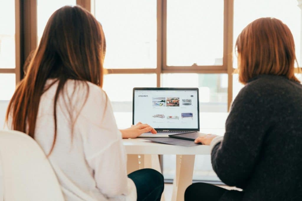 What UK Competition Website Owners Need to Know Before Launching Two people sit at a white table looking at a laptop displaying a product gallery on a bright windowed background.