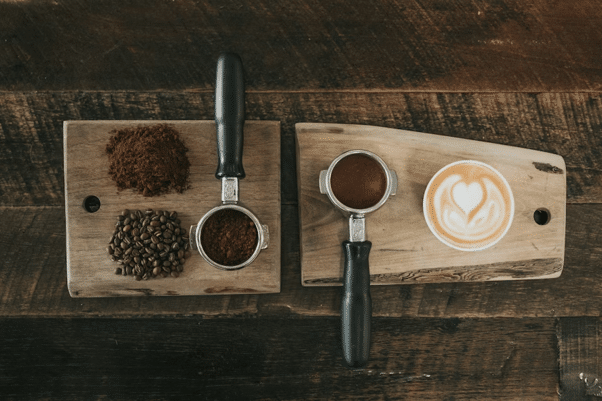 Understanding the Different Types of Coffee Capsules Two wooden boards hold espresso portafilters, ground coffee, and a cup of latte with latte art on a dark wooden table.