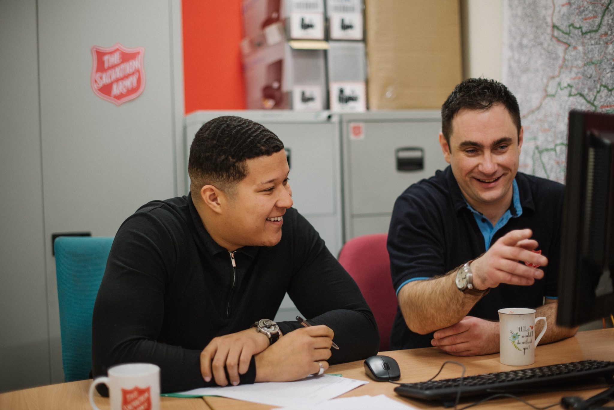 Two men sit at a desk in an office, smiling while looking at a computer screen with coffee mugs nearby.
