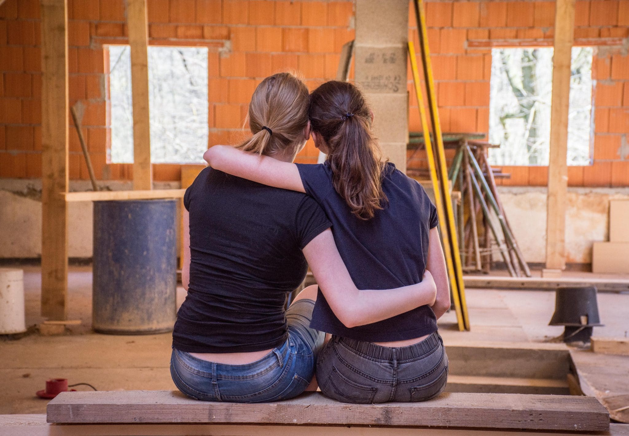Two people sit close with arms around each other, facing a construction site with orange brick walls and tools in the background, conveying support or friendship.