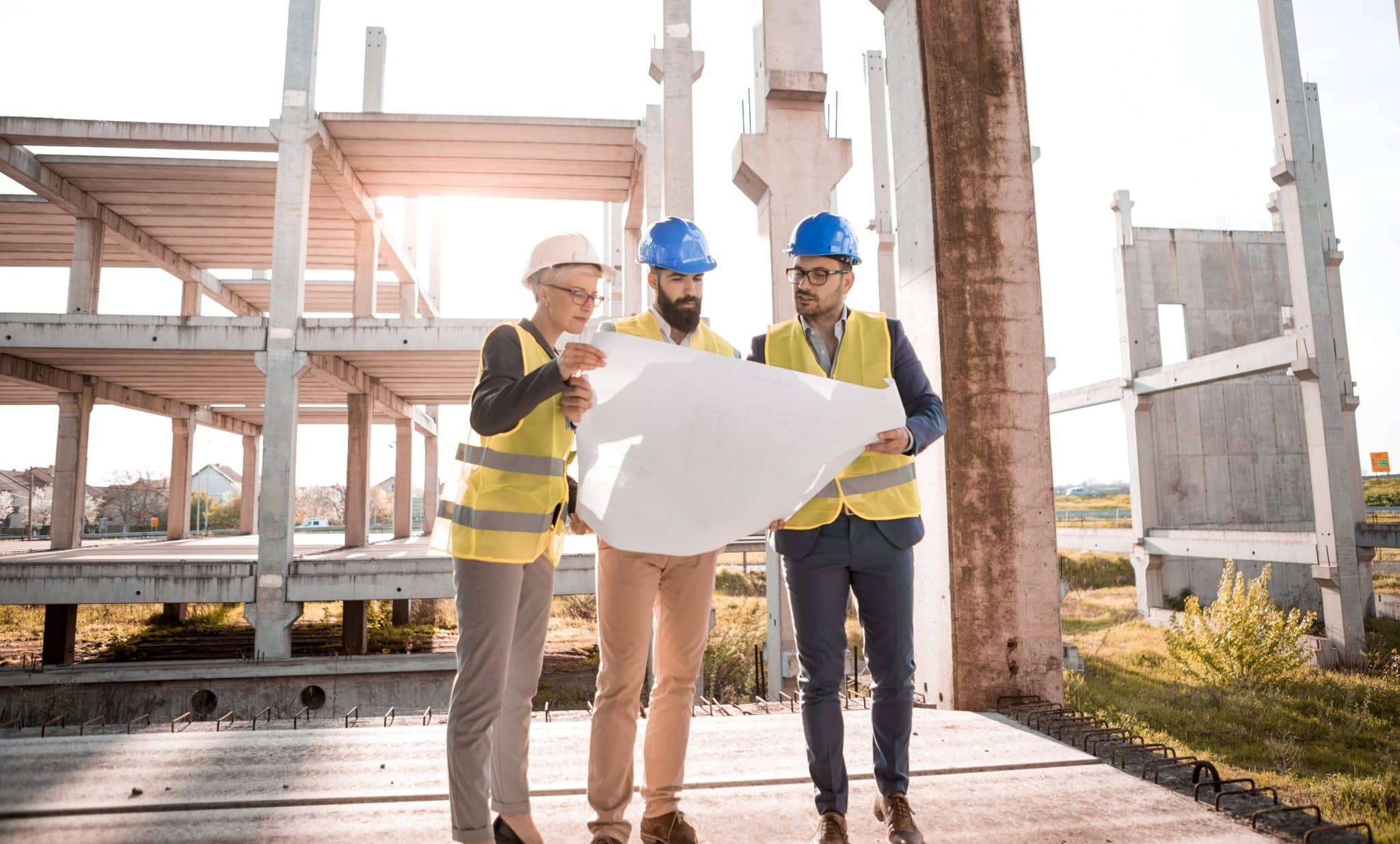 Three construction workers in safety gear review blueprints at a building site under a concrete frame sun glare to the left