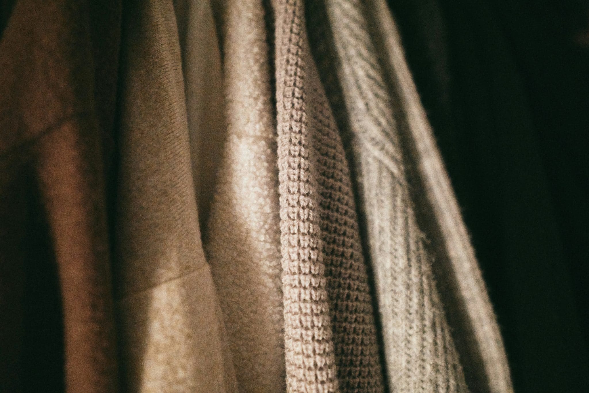 Close-up of folded neutral-colored fabrics or scarves hanging in a rack, showing varied textures and knits in warm browns and beiges.
