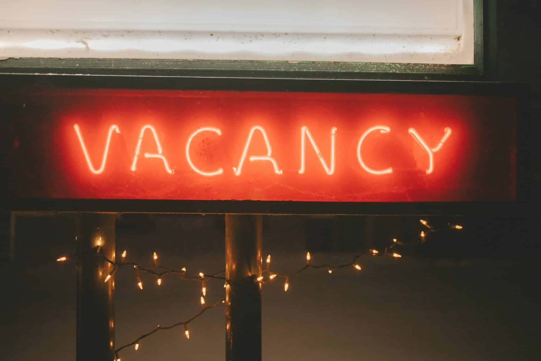 Red neon 'VACANCY' sign in a storefront window with string lights hanging outside.