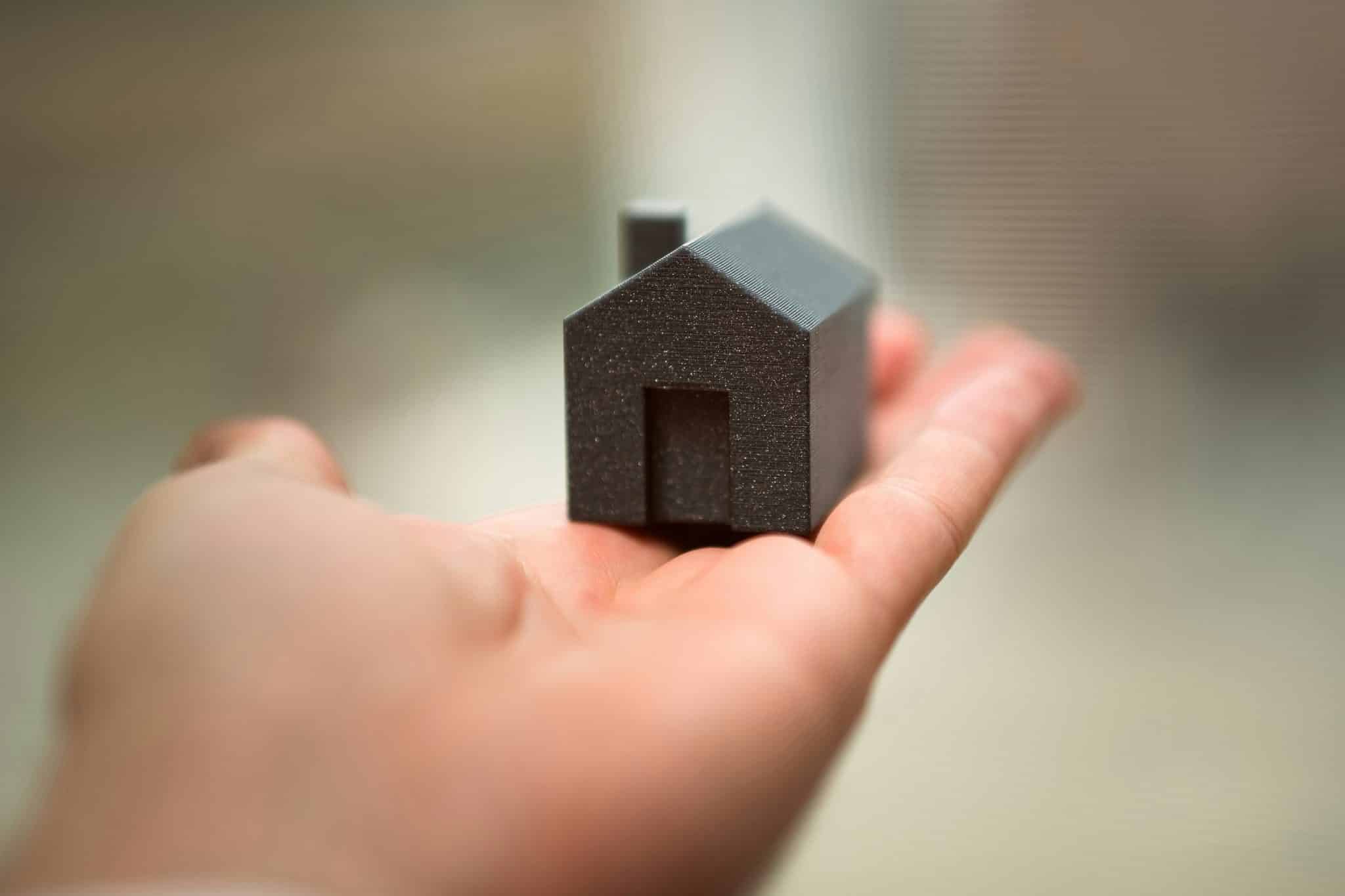 Small black house-shaped model held between two fingers, close-up hand view, likely a miniature or 3D-printed prototype.