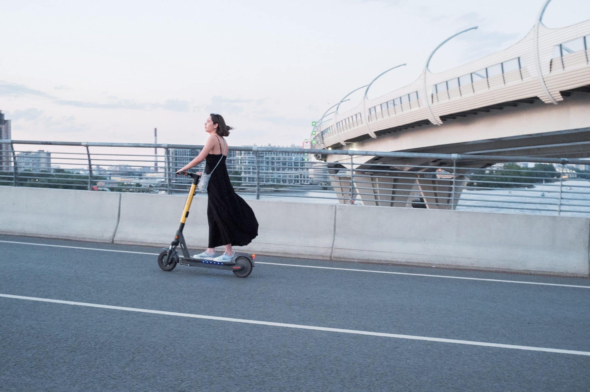 Woman wearing a black dress rides a yellow electric scooter on a bridge with railing and city skyline in the background for a calm urban scene