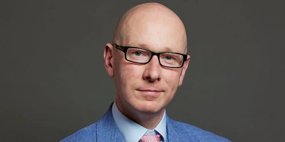 Professional headshot of a bald man wearing black-framed glasses, a light blue dress shirt, pink tie, and blue suit jacket against a gray backdrop
