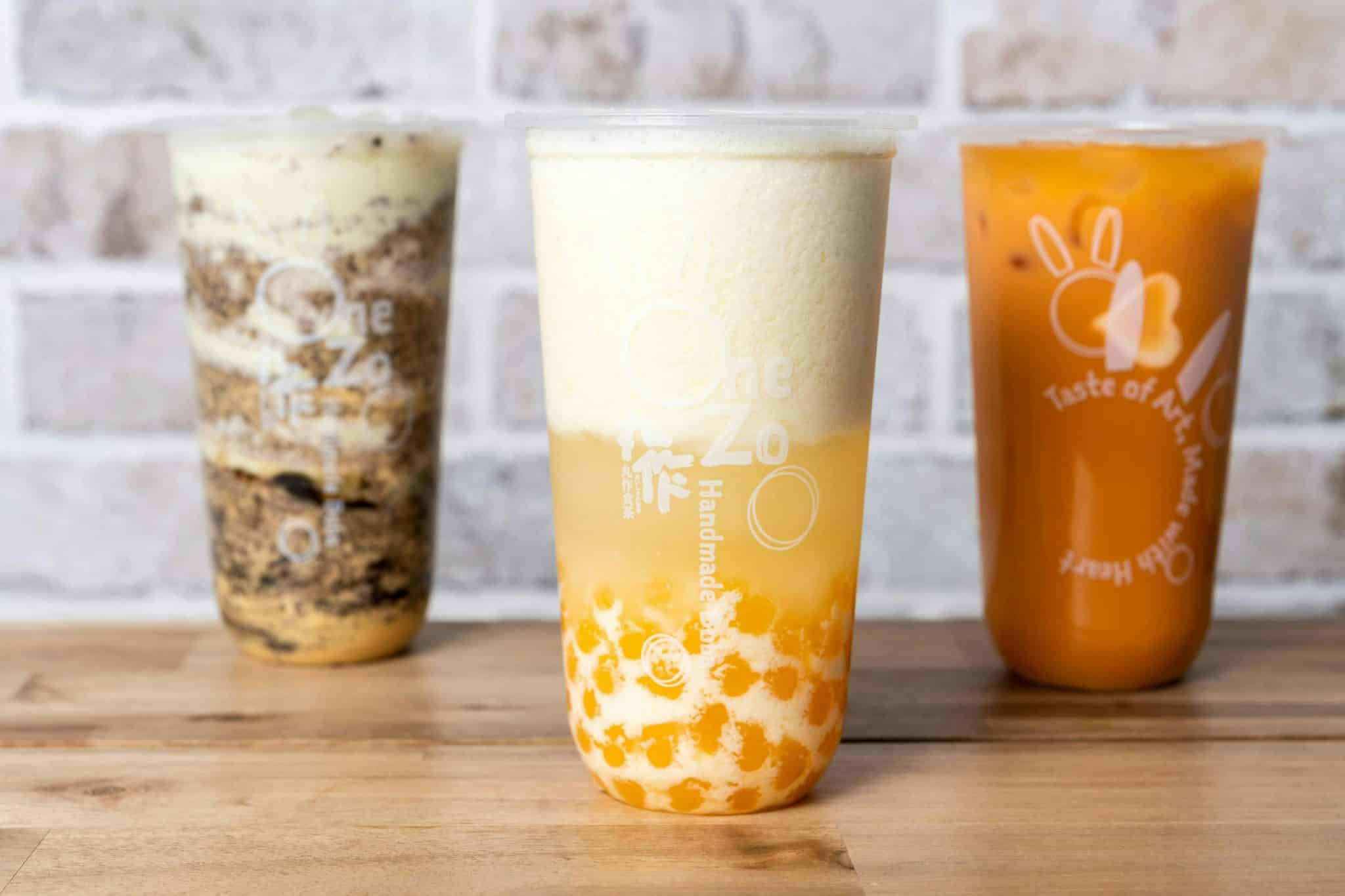 Three iced beverages in clear cups on a wooden surface; left chocolatey with layers, center creamy pale yellow, right bright orange with a white logo and graphics.