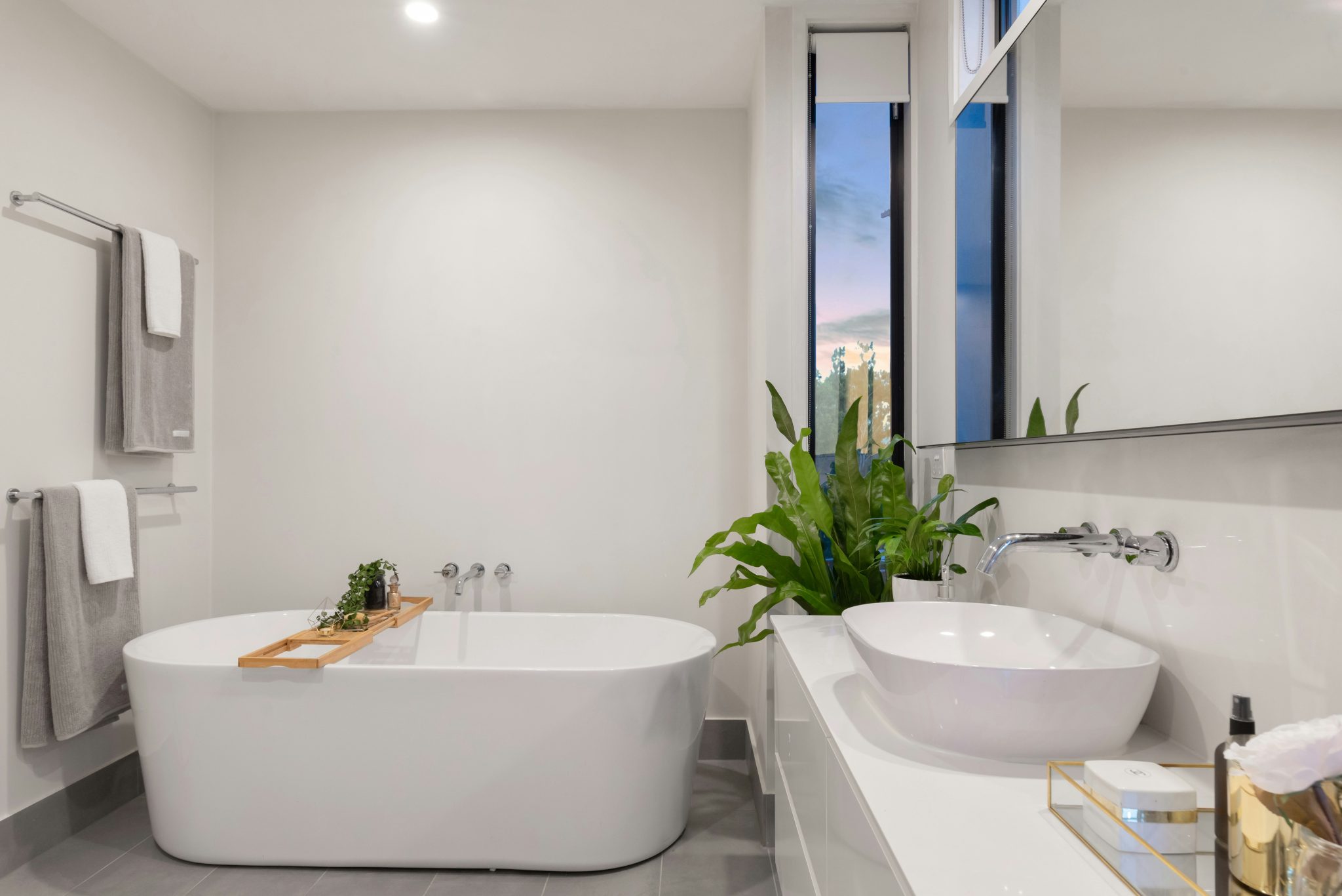 Modern bathroom with a white freestanding bathtub, double sink vanity, and green plants by a tall window; gray tiled floor and towels on a rack on the left