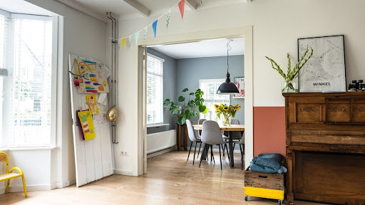 How to Choose the Right Wooden Flooring for Your Home Bright open-plan room with a doorway to a blue dining area; colorful bunting, potted plants, and a vintage piano against a two-tone wall.