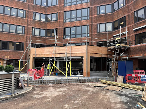 Construction entrance under renovation: scaffolding around a brick building, workers in hard hats, yellow braces, and orange safety barriers with wooden framing.