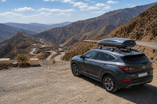 Gray SUV with a roof cargo box parked on a dirt overlook, overlooking a winding mountain road and rugged canyon scenery inside a blue sky. You could describe the scene as a rugged mountainous drive setup.