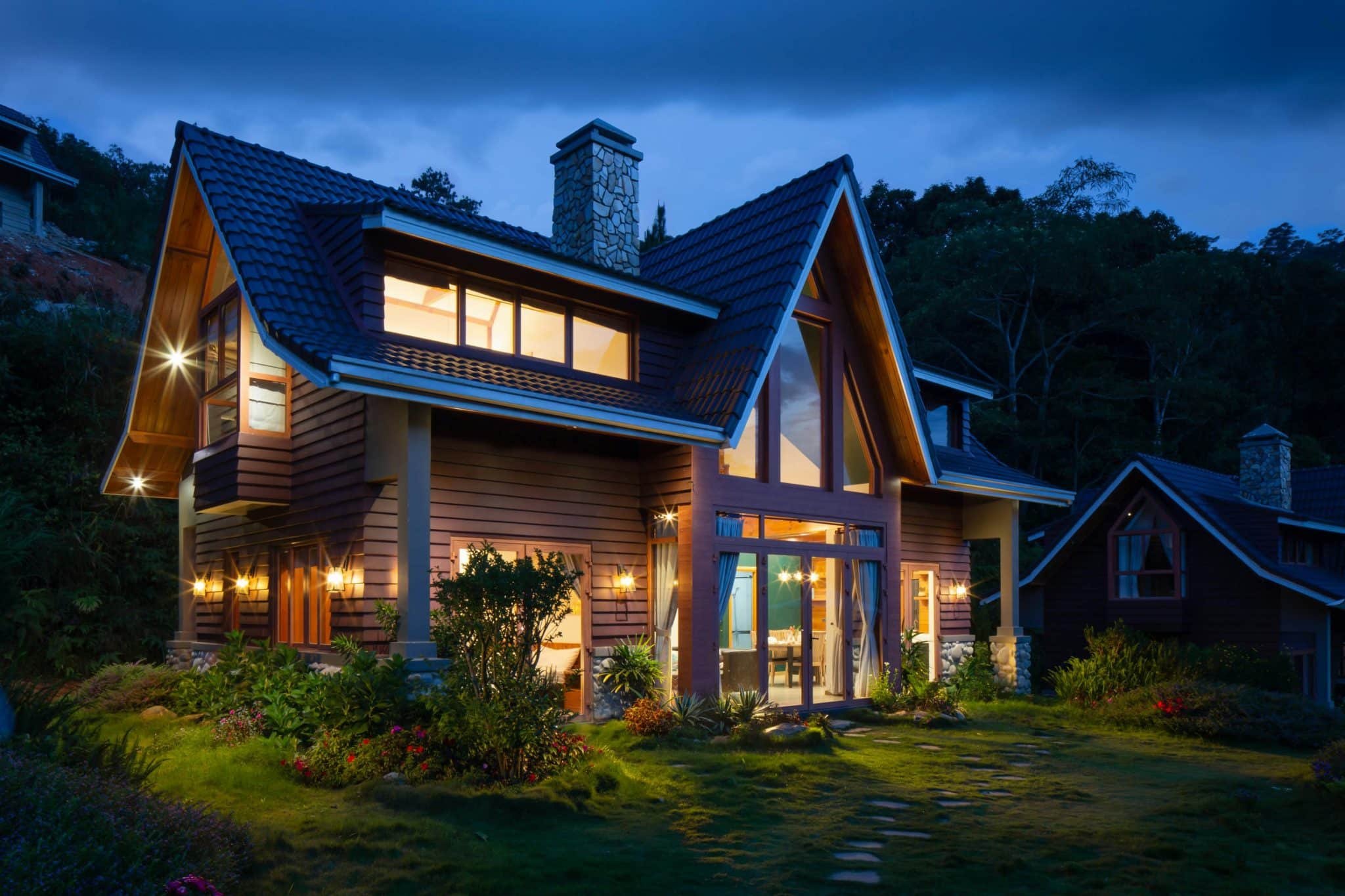 Modern two-story house with large glass windows glowing warmly at dusk, surrounded by a garden.