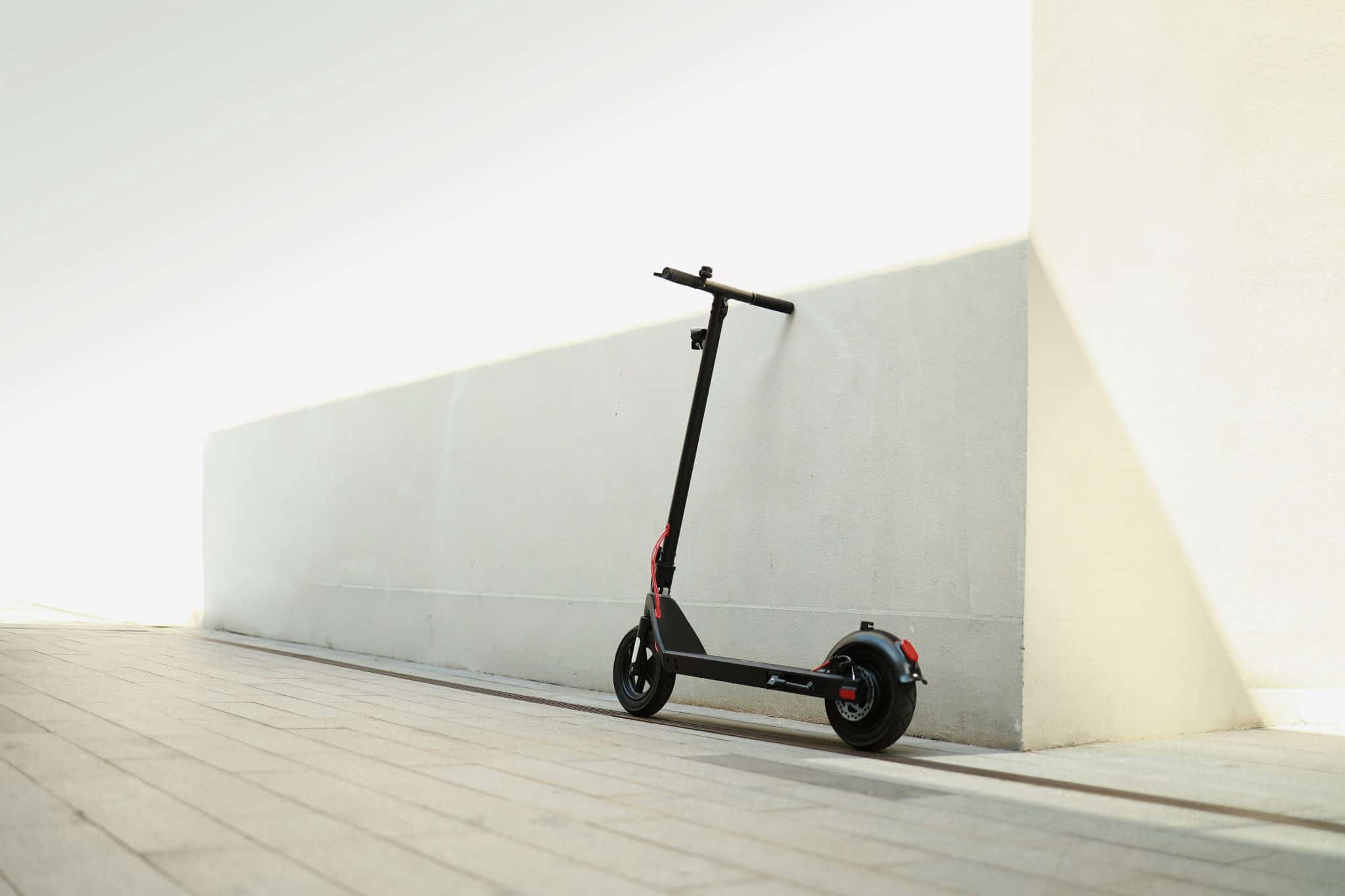 Electric scooter leaning against a pale wall in bright sunlight, handlebars at mid-height and red wiring visible near the stem.