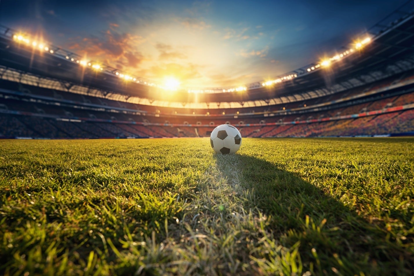 Soccer ball centered on sunlit grass inside a large stadium with stands in the background.