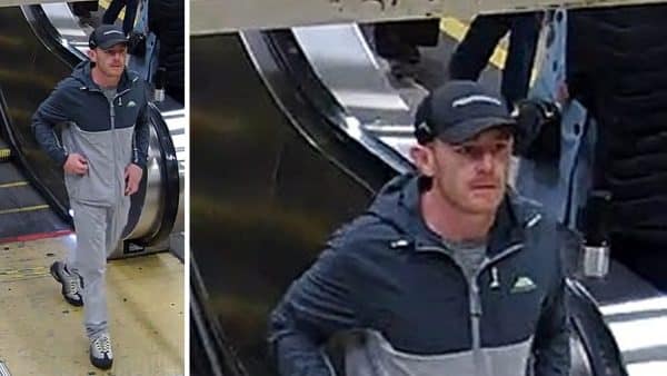 Man in a gray and navy two-tone jacket and black cap steps off an escalator in a transit station.