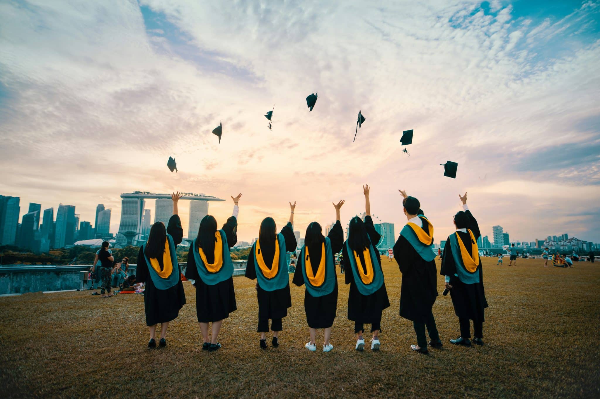 Graduates in black gowns toss their mortarboards into the air at sunset, with a city skyline in the background.