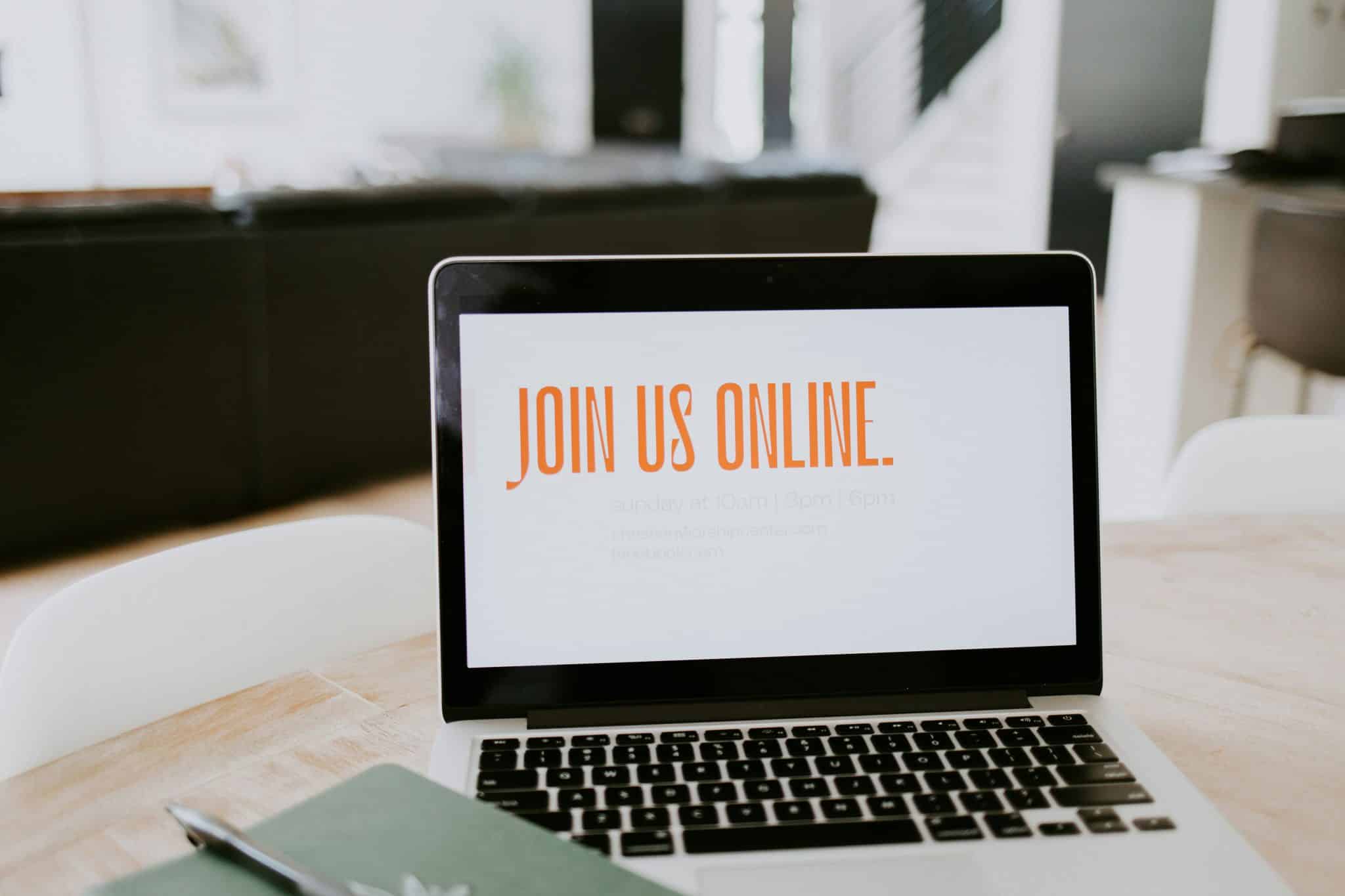 Laptop on a desk showing the phrase 'JOIN US ONLINE' in orange on the screen (background blurred).